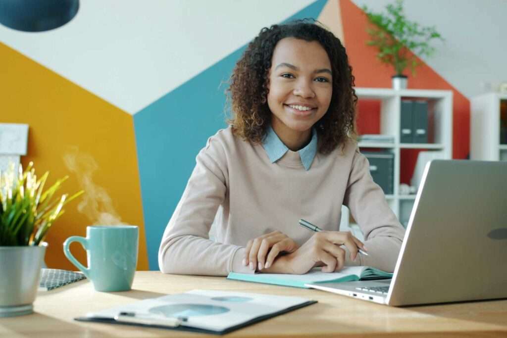 Junge Frau lächelt am Schreibtisch mit Laptop und Kaffee
